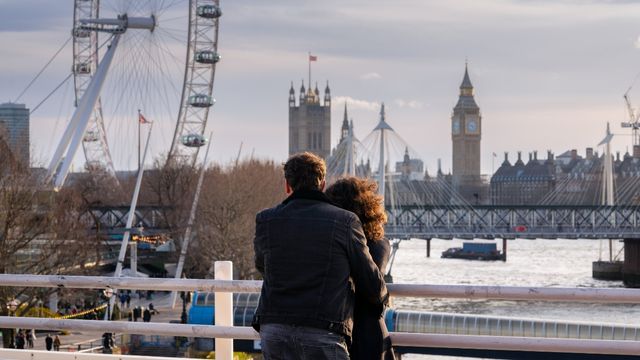 A couple stands together by the river thames looking at the london eye and tower bridge.
