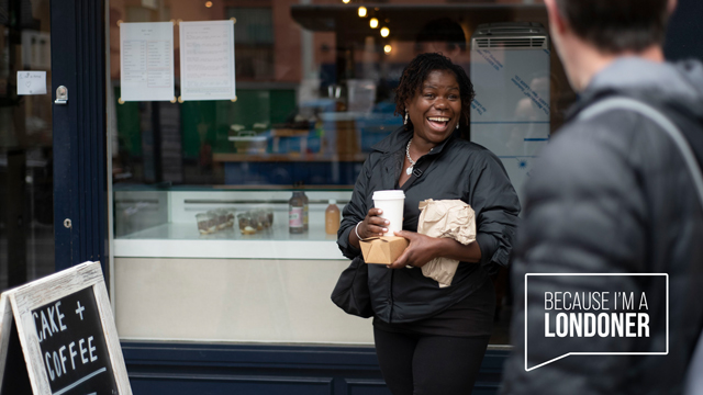 A lady stands in front of a shopfront with a takeaway coffee cup and food wrapped in a brown paper bag, she's smiling as a man walks past in the foreground. A logo worded "Because I'm a Londoner" is overlaid on the image, inside a speech bubble.