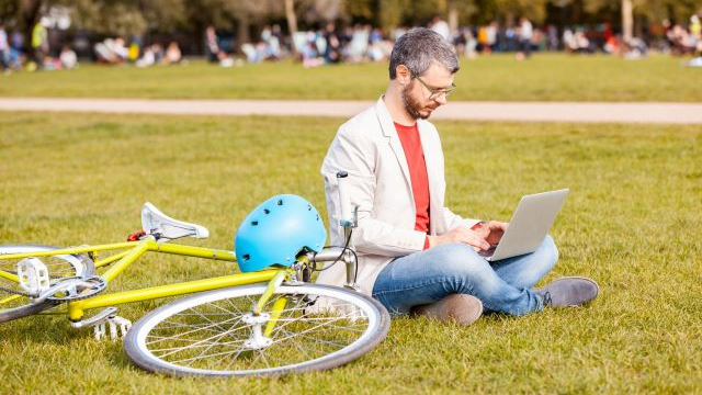 Man working on his laptop sitting on the grass at a park next to his bike