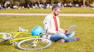 Man working on his laptop sitting on the grass at a park next to his bike