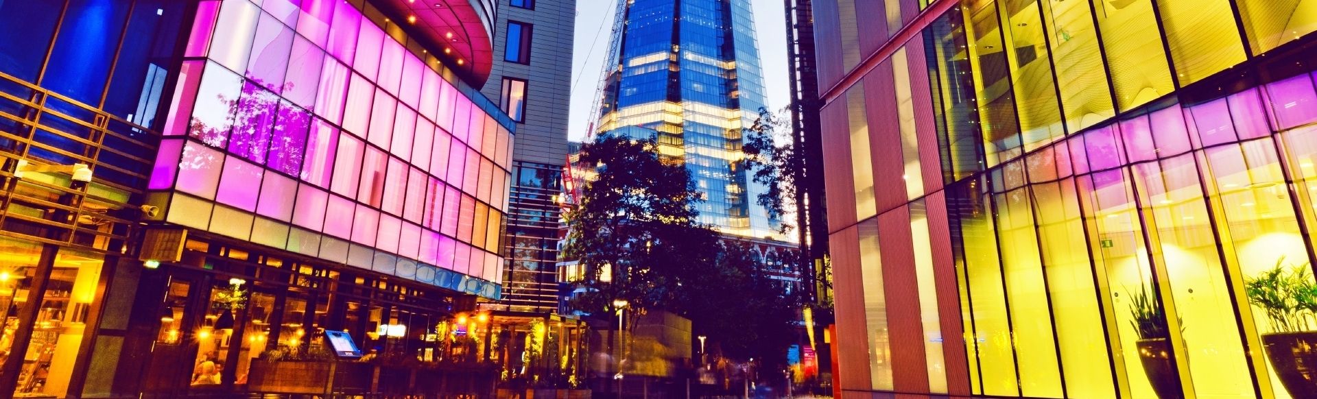 The Shard in the centre of a walkway. There are buildings with pink, blue and yellow glass panels on both sides of the path.