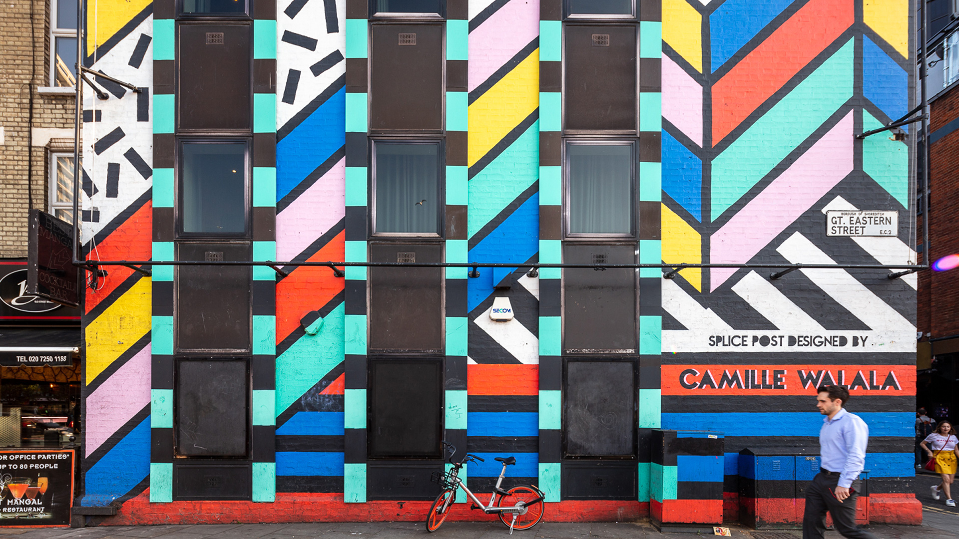 Man walking past the facade of a building painted with vibrant colours by an artist Camille Walala whose name is written on the bottom right corner of the building 