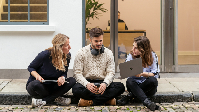 Three colleagues sitting on a step outside of an office looking at something on a laptop