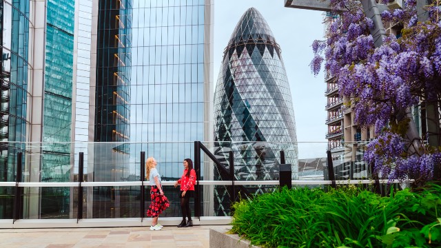Two women stood chatting in front of 120 Fenchurch Street, beside other glass skyscrapers and hanging purple flowers.