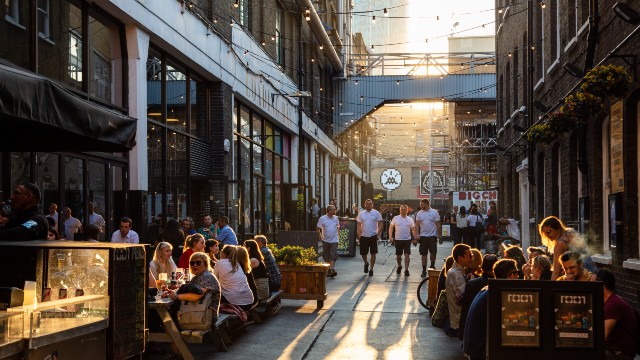 People walking down a large alley with people sat outside various restaurants either side as the evening sun streams down the middle.