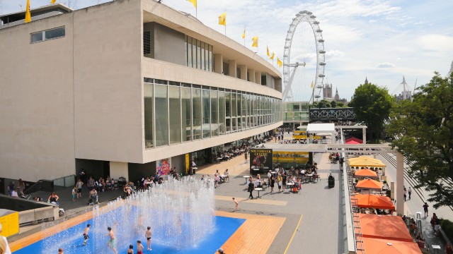 People paddling in a blue pool with a building next to it and the London Eye in the distance.