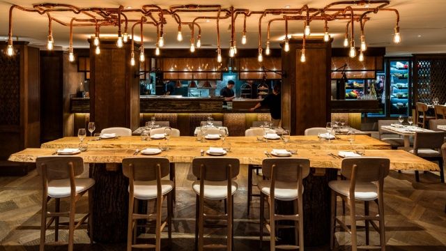 Decked wooden dining table with 10 seats, with a designer lamp made out of copper pipes shining from above.