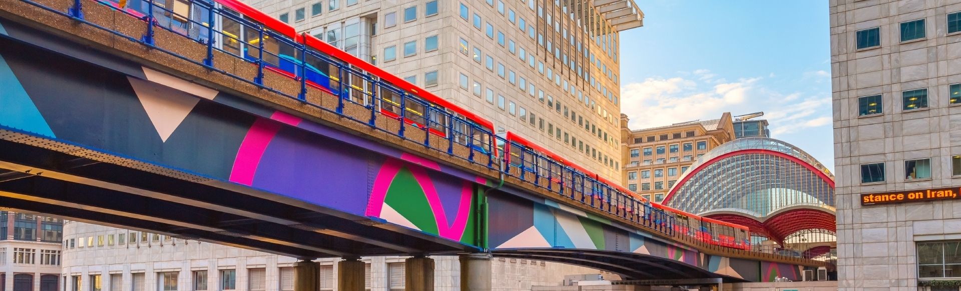 Colourful bridge in Greenwich, Canary Wharf DLR station, with a purple, pink, green and blue pattern on the side. The bridge carries a public transport train and runs in between two buildings.