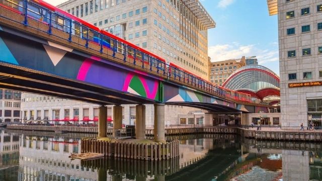 Colourful bridge in Greenwich, Canary Wharf DLR station, with a purple, pink, green and blue pattern on the side. The bridge carries a public transport train and runs in between two buildings.