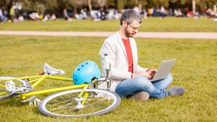 Man sitting on the grass at a park working on his laptop next to his bike.