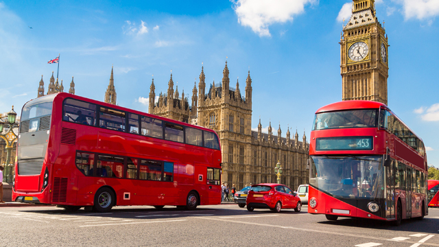Two red doubledecker buses driving on the main street in front of the Houses of Parliament on a sunny day.