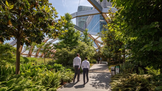Two men in dark trousers and white shirts walk along a path lined by green plants underneath a glass canopy.