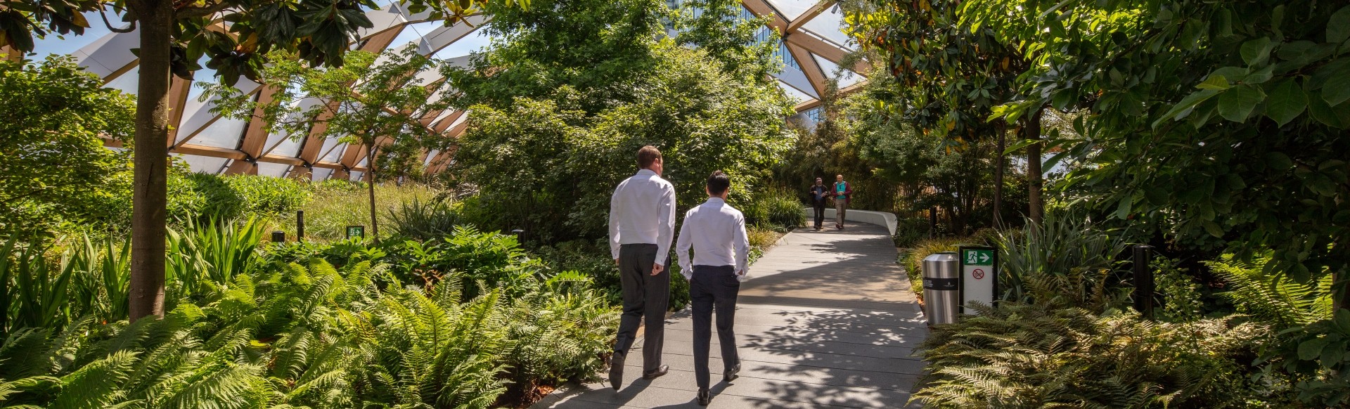 Two men in dark trousers and white shirts walk along a path lined by green plants underneath a glass canopy.