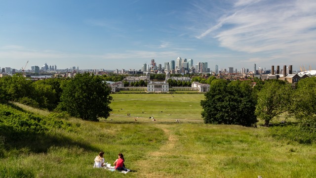 Two people sit on a picnic blanket and take in the view from the top of the hill in Greenwich Park with the London skyline in the distance.