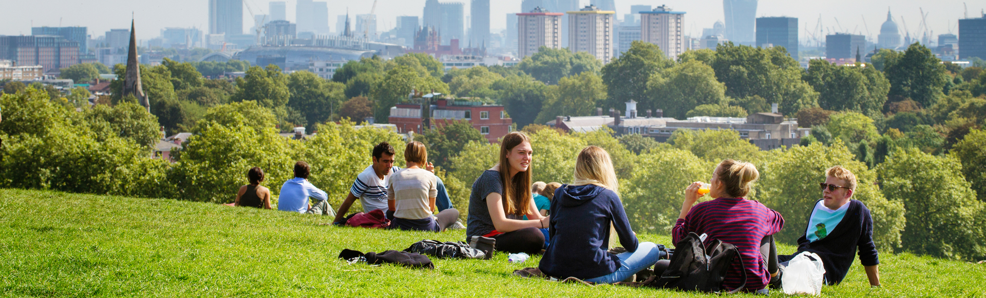 A group of people sitting in a green and sunny park at Hampstead Heath with the London skyline in the background.