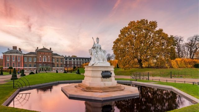 Fountain with a white statue in the middle in Kensington Palace gardens, with a rosy sunset sky in the background, reflecting in the water.