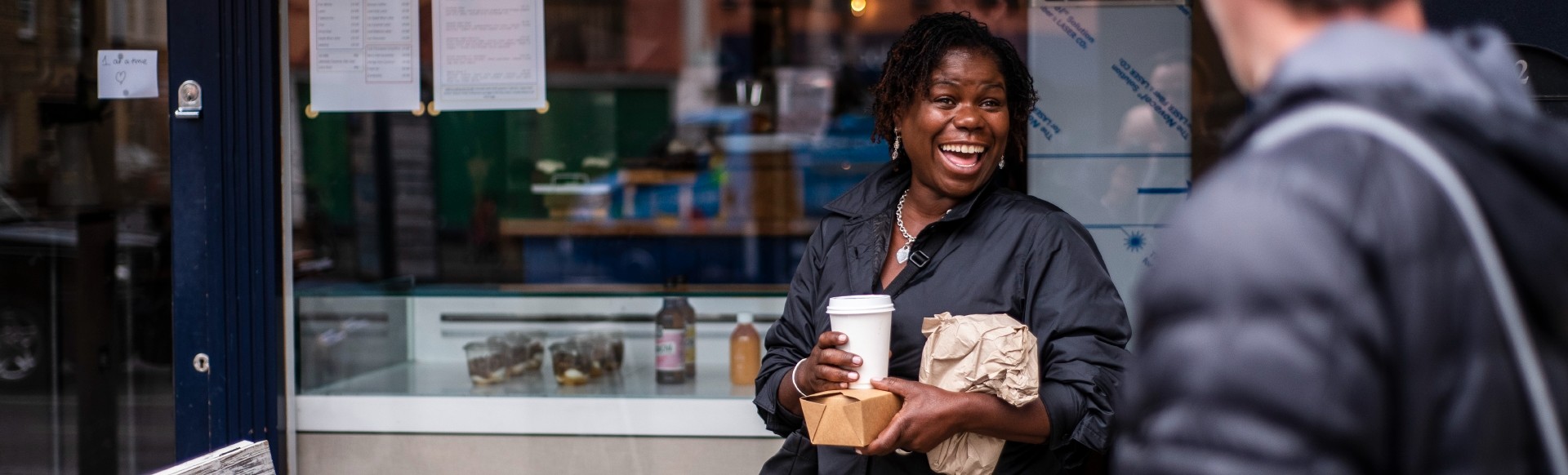 A woman smiles while standing outside of a cafe with a coffee cup in hand.
