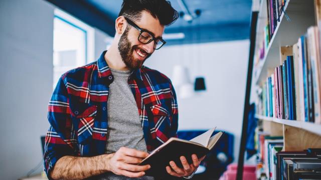 A bearded male student in a check shirt looks at a book in a library.