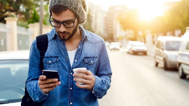 A young bearded man is walking on the street, looking at his mobile phone, as the sun rises behind him.
