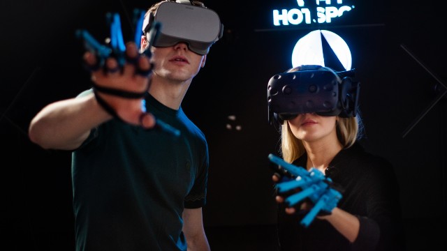 Two young people are standing next to each other in a black room wearing VR helmets and gloves.