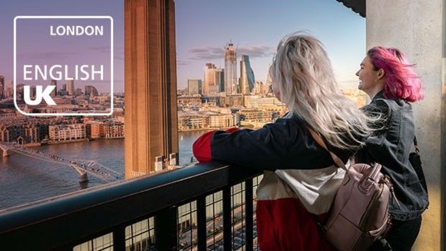 Two young women are standing on a outdoor terrace, facing London's skyline on a sunny day.
