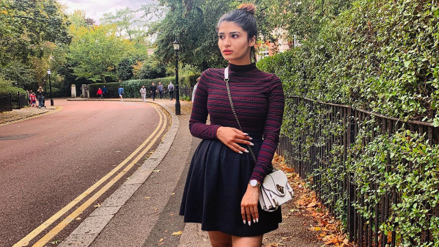 Smriti is standing by a leafy hedge on a curbed road.