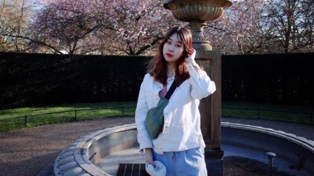 A young woman wearing a white hoodie is standing in front of a fountain and cherry blossomed trees on a sunny day.