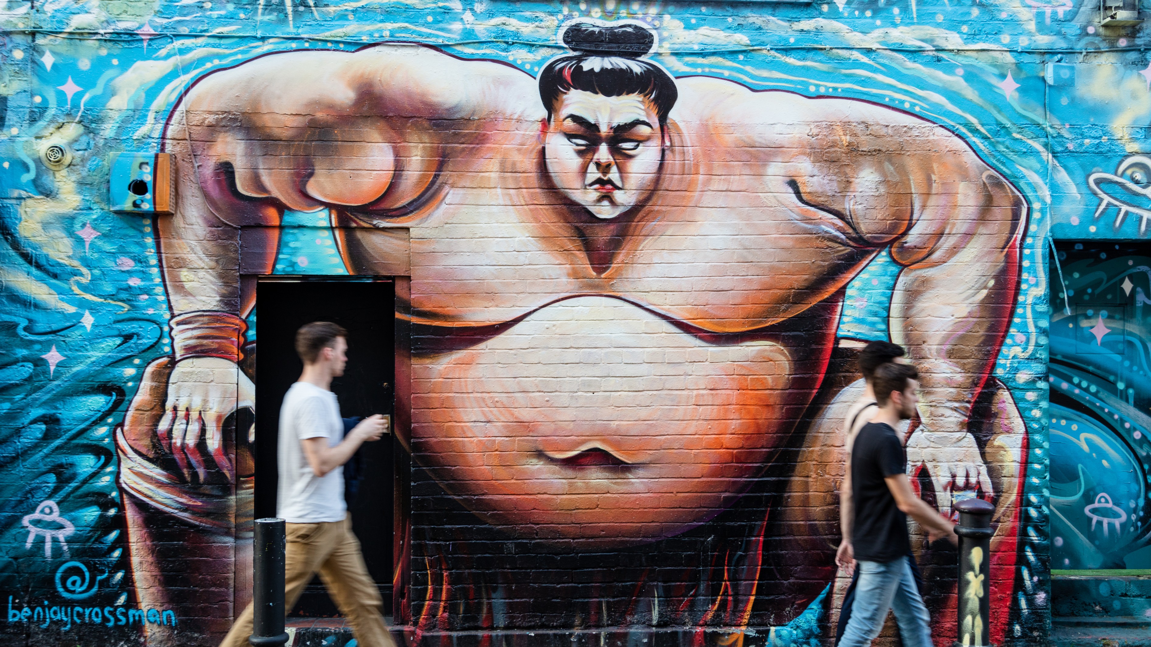 Graffiti mural of a sumo wrestler against a blue background on a brick wall in Spitalfields.