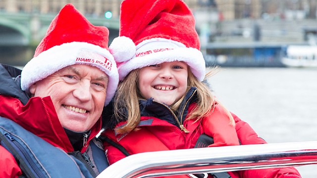 Ein Mann und ein Kind mit Weihnachtsmannmützen lächeln, während sie eine Fahrt mit dem Speedboot auf der Themse genießen.