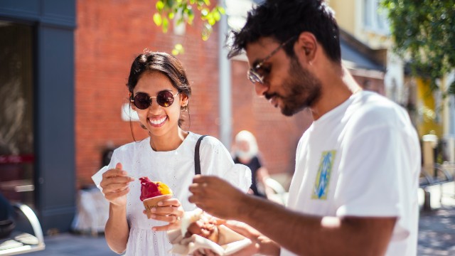 A man and a woman wearing sunglasses smile while tucking into takeaway food in the sunshine as they explore Brixton. 