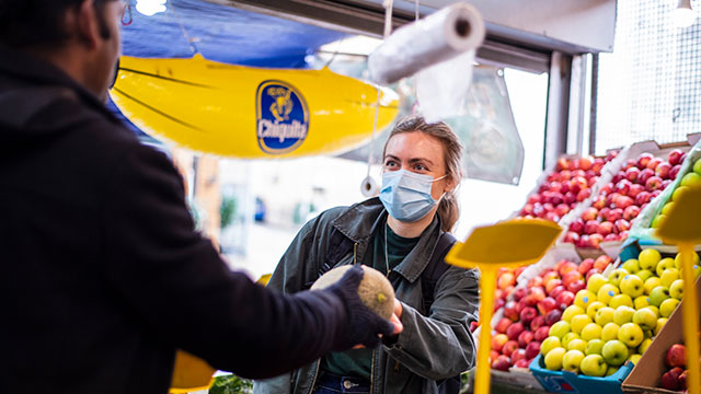 A woman wearing a mask hands over a melon to a customer at a fruit stall in a market.
