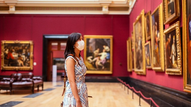A person wearing a mask admires a wall filled with paintings at the National Gallery.