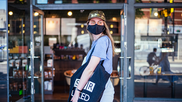 A woman, wearing a face covering and carrying a bag, waits to go inside a shop.