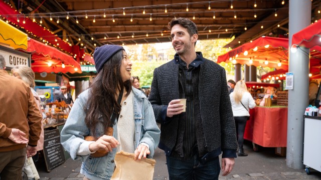 Two people smile while walking through the stalls of Borough Market illuminated with festoon lights 