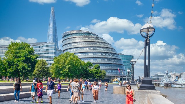 A crowd of people stroll along the walkway next to the river Thames on a sunny day with The Shard and City Hall in the background