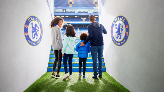 Une famille de quatre personnes traverse le tunnel du Chelsea Football Club en direction du stade baigné de lumière, semblant très enthousiaste.