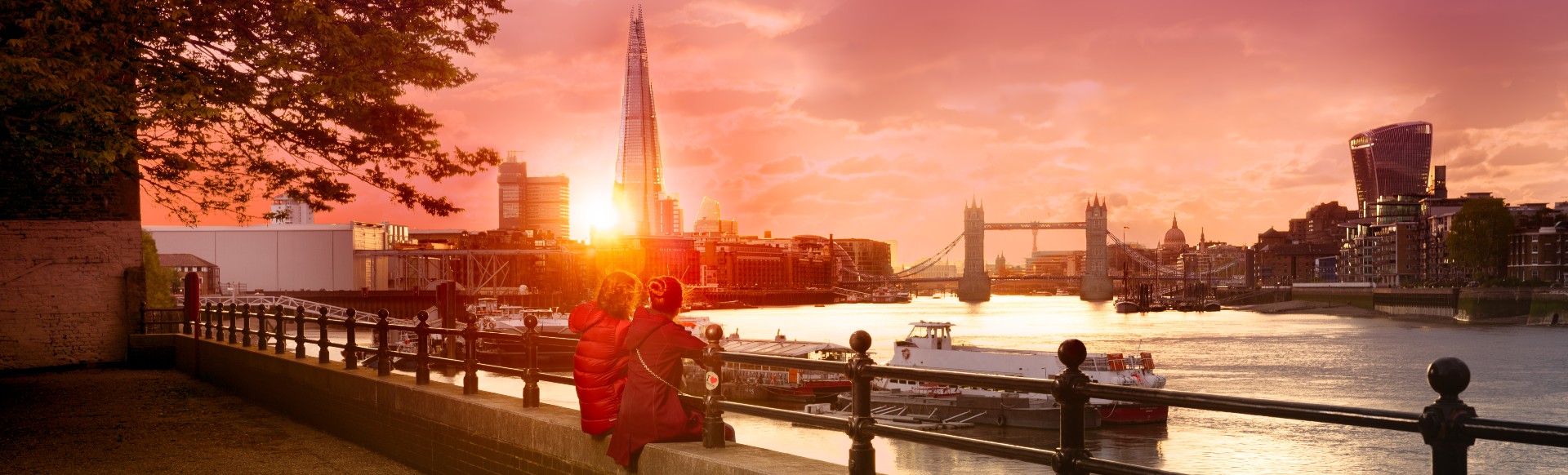Let's Do London. Credit: Antoine Buchet. Image courtesy of Antoine Buchet. Two people sit on the banks of the river Thames to watch the sun set over the London skyline including The Shard and Tower Bridge.