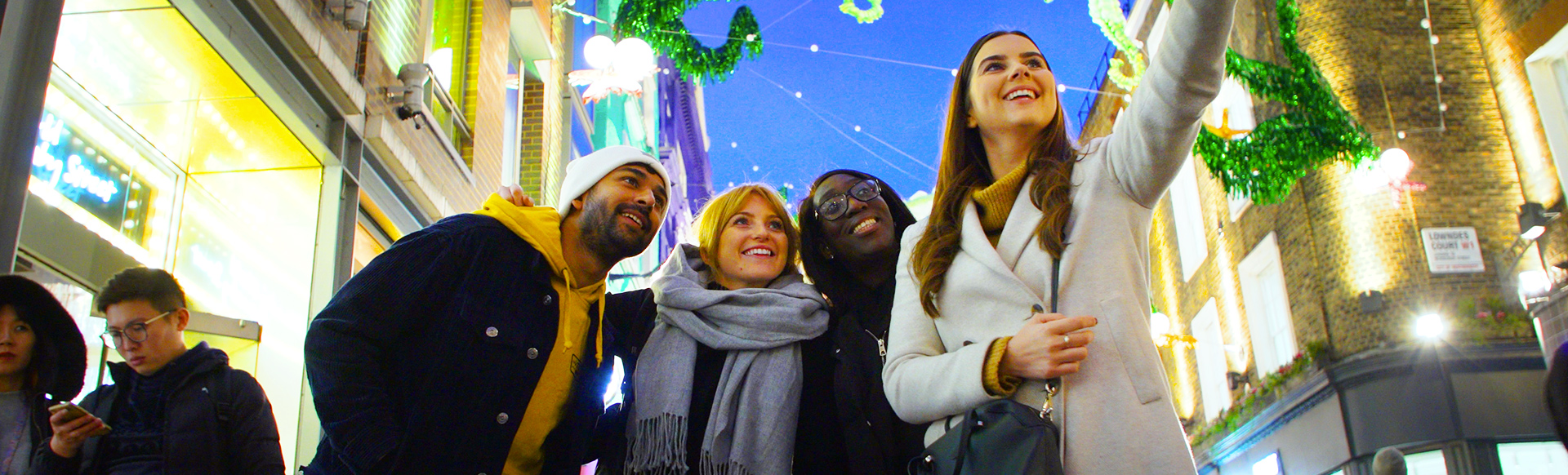 Let's do London. Christmas lights in Carnaby. Image courtesy of TMW Unlimited. A group of four young adults takes a selfie in front of the Christmas lights on Carnaby Street, with green seahorse decorations suspended between buildings.