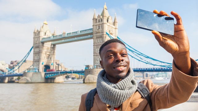 Un homme lève son téléphone pour prendre un selfie avec le Tower Bridge derrière lui, par une journée ensoleillée.