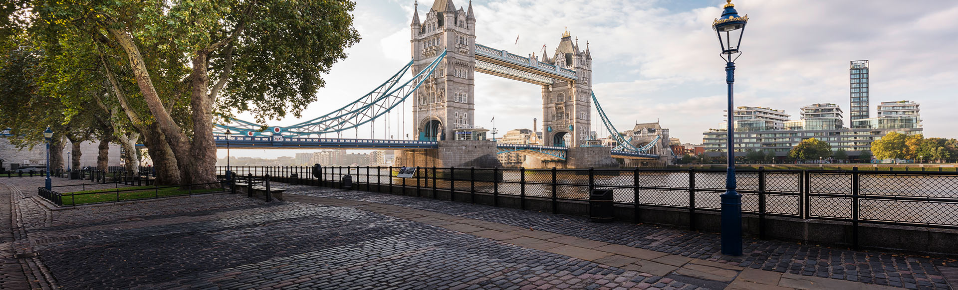 A view of Tower Bridge with a cobbled walkway, lamp-post and tree in the foreground.