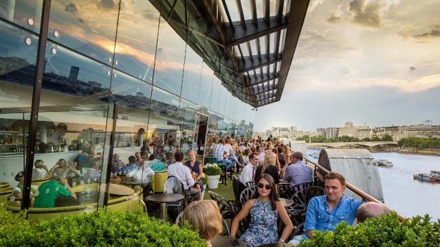An outdoor rooftop overlooking the London skyline, with people eating and drinking in the hazy sunshine.