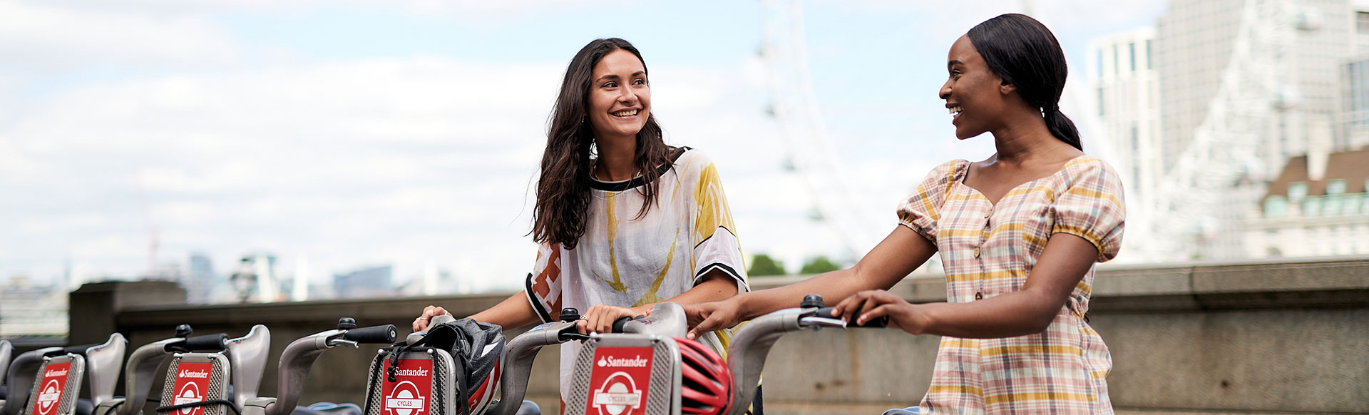 Two ladies in short-sleeved clothing smile at each other while holding a Santander Cycles bicycle each, with the London Eye in the background.