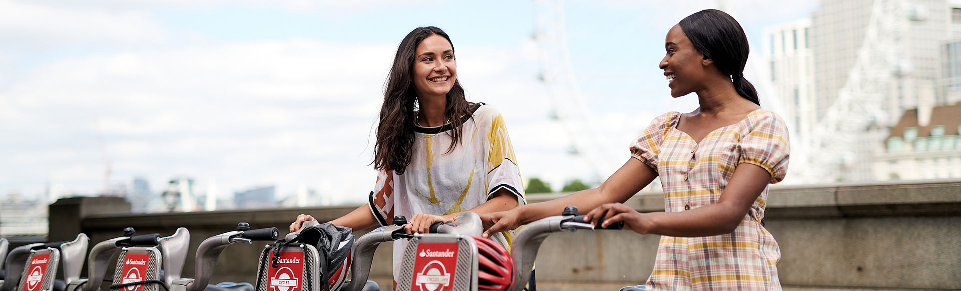 Explore London by bicycle during a staycation © London & Partners/Sam McElwee. Two ladies in short-sleeved clothing smile at each other while holding a Santander Cycles bicycle each, with the London Eye in the background.