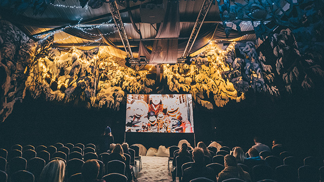 People seated in rows watching The Muppets Christmas Carol on a cinema screen, surrounded by foliage dusted in snow, and with snow on the ground.