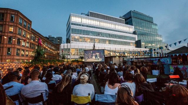 People sitting on the rooftop at Queen of Hoxton at dusk, watching a film on the pop-up screen, with skyscrapers in the background.