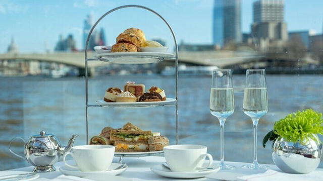 An afternoon tea spread on a table including two glasses of champagne and a tiered tray of cakes, scones and sandwiches with the river Thames in the background