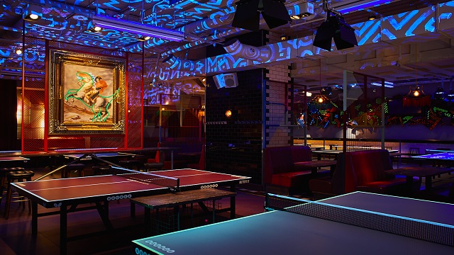 A dark room with club-style lighting and ping pong tables laid out