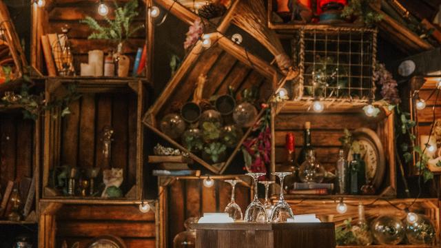 Wooden boxes arranged as shelves with books, glasses, plants and bottles of alcohol on display in a darkly lit room.