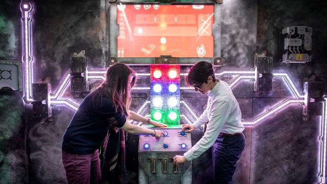 Two people stand by a control board with multi-coloured lights and buttons in a london escape room.