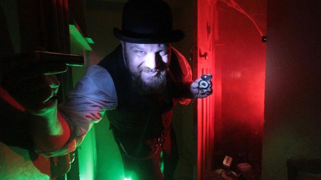 A man in a black bowler hat stands under red and green light holding an escape room clue.